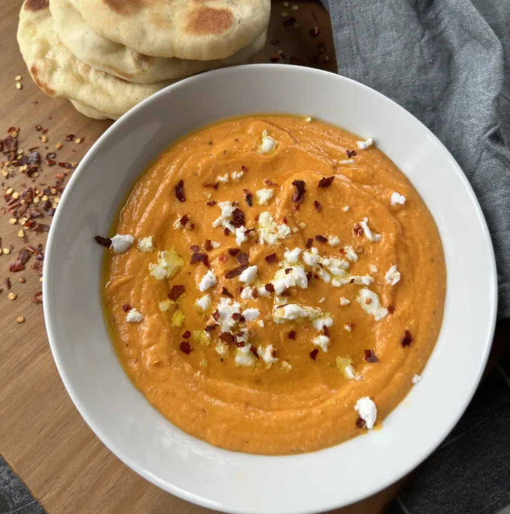 Creamy feta and roasted red pepper dip with an orange colour, topped with some more vegan feta, chilli flakes and served with fresh flatbreads