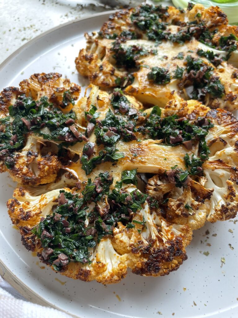 close up shot of Two Greek style seasoned roasted cauliflower steaks on a plate topped with parsley and kalamata olives