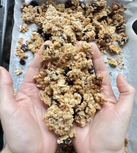 Hands holding a handful of cherry and vanilla granola from the baking tray below it with the rest of the granola on