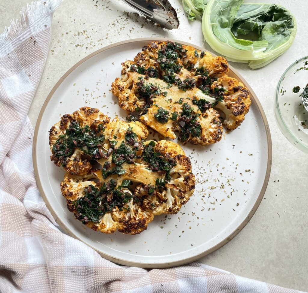 Two Greek style seasoned roasted cauliflower steaks on a plate topped with parsley and kalamata olives